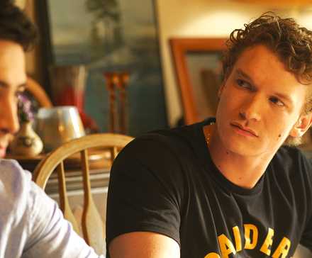 Connor Storrie and Hudson Williams looking tense sitting at a table in the Heated Rivalry finale.