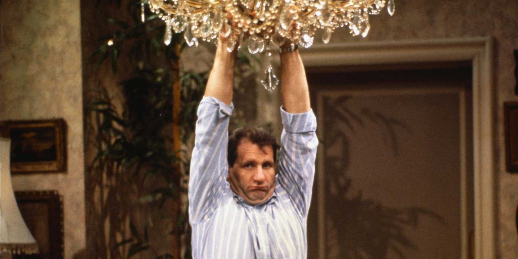 Ed O'Neill as Al Bundy hangs from a chandelier with an amusingly confused look in Married...With Children.