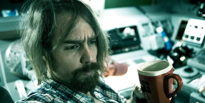Sam Rockwell as Sam Bell in Moon, with long hair and an unkempt beard, looking slightly ahead and to the right while holding a brown coffee mug in his right hand