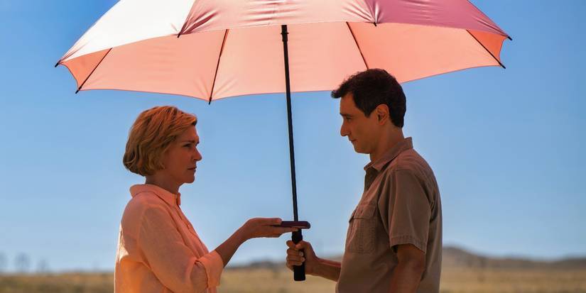 Rhea Seehorn holds an object out to Carlos-Manuel Vesga under an umbrella in the Pluribus finale.