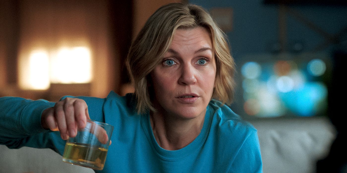 Rhea Seehorn dengan kemeja biru, duduk dan memegang minuman sambil melihat ke depan di Pluribus.