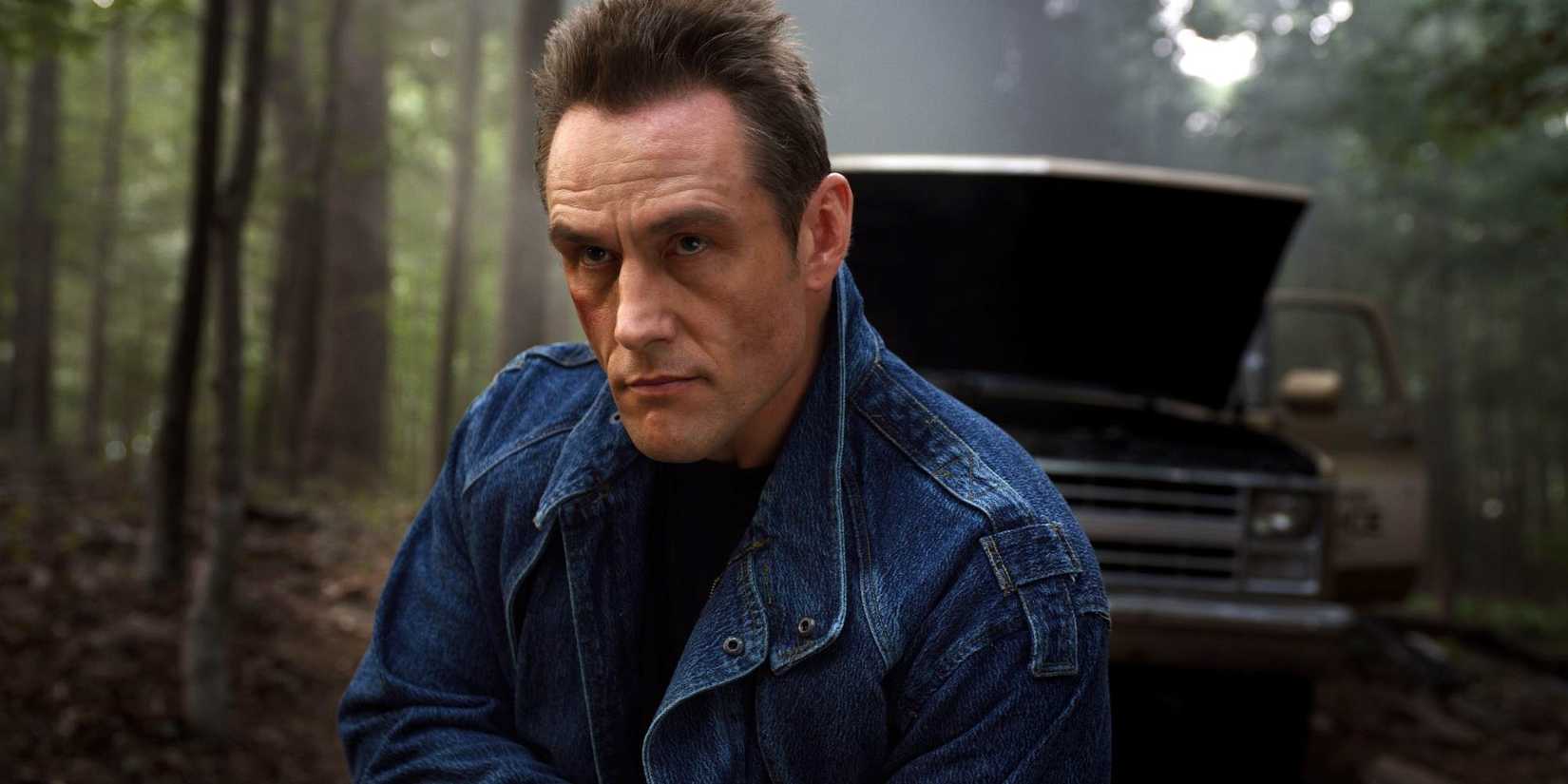 Andrey Ivchenko as Grigori in a denim jacket with a broken vehicle behind outdoors in Stranger Things.