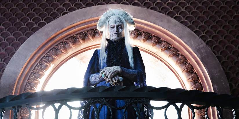 An ancient-looking Dracula stands in front of a window, over a banister, looking down.