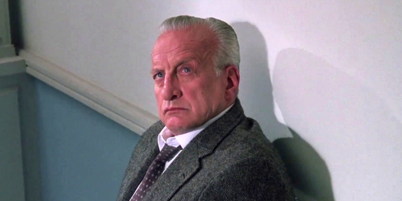 George C. Scott seated against a wall, wearing a suit jacket and tie in The Exorcist III