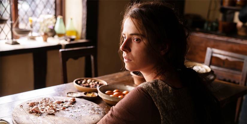 Agnes sits at a table with herbs and ingredients in front of her, looking over her shoulder.