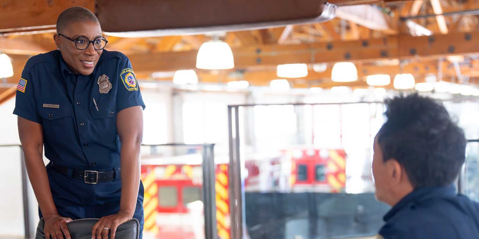 Hen standing behind a chair and talking to a seated Chimney in 9-1-1 Season 9