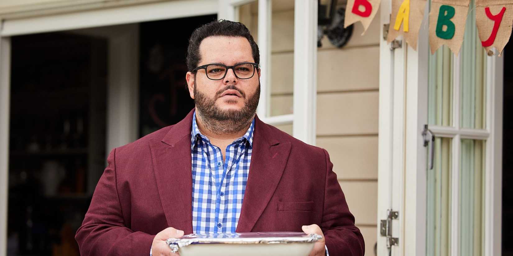 Josh Gad as Gary carries food at an outdoor party with a banner that partially reads "baby" behind in Wolf Like Me. 