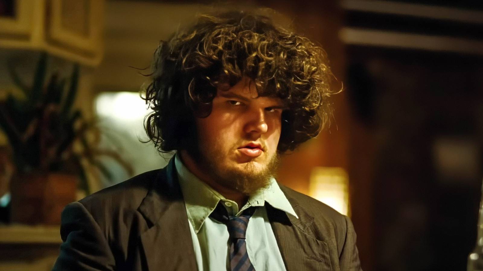 Aidan Delbis sits at a table in a suit, looking skeptical in Bugonia