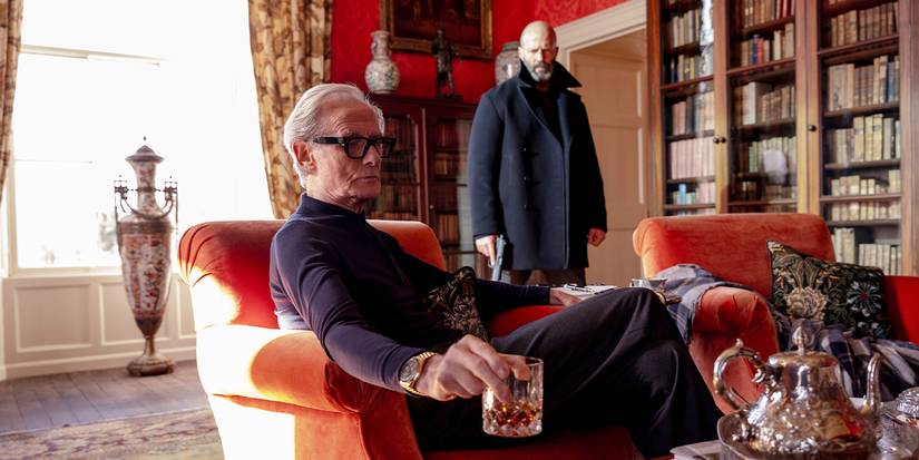 Mason stands menacingly behind Bill Nighy's character, who is sitting in an armchair with a drink.