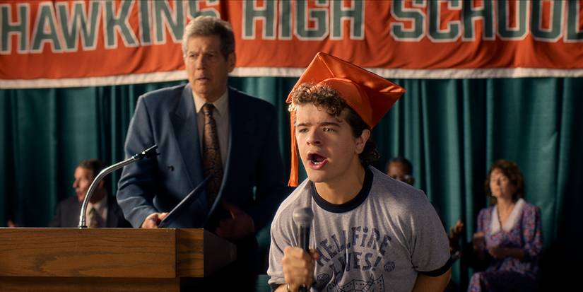 Dustin talking into the mic wearing a "Hellfire Lives" shirt at graduation in Stranger Things.