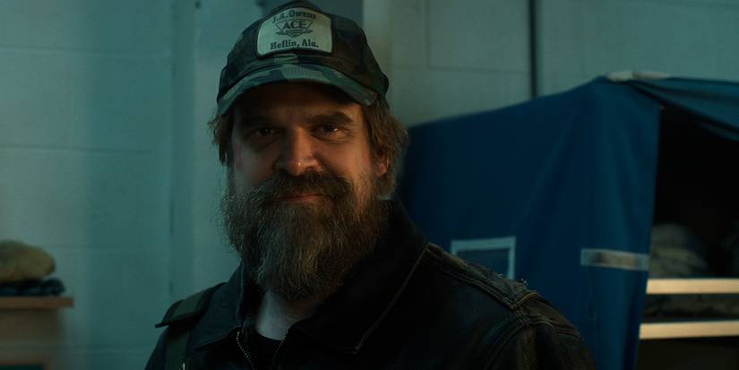 Hopper with a beard and baseball cap smiling in Stranger Things.