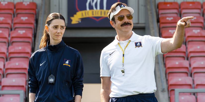 Ted Lasso pointing while wearing a visor and sunglasses, a woman beside him.