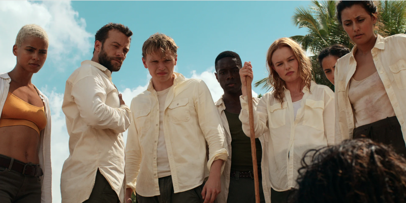 The I-Land cast standing together looking down seriously at someone on the ground under a bright sky.