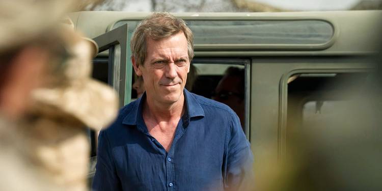 Roper standing in front of a military vehicle and smiling in The Night Manager Season 1