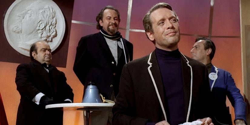 Number Six smugly smiling as three men stand behind him in The Prisoner