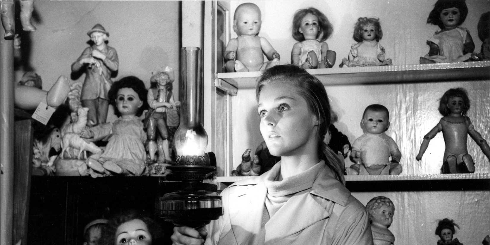 Carol Lynley surrounded by dolls on shelves with a lantern in her hands in Bunny Lake Is Missing