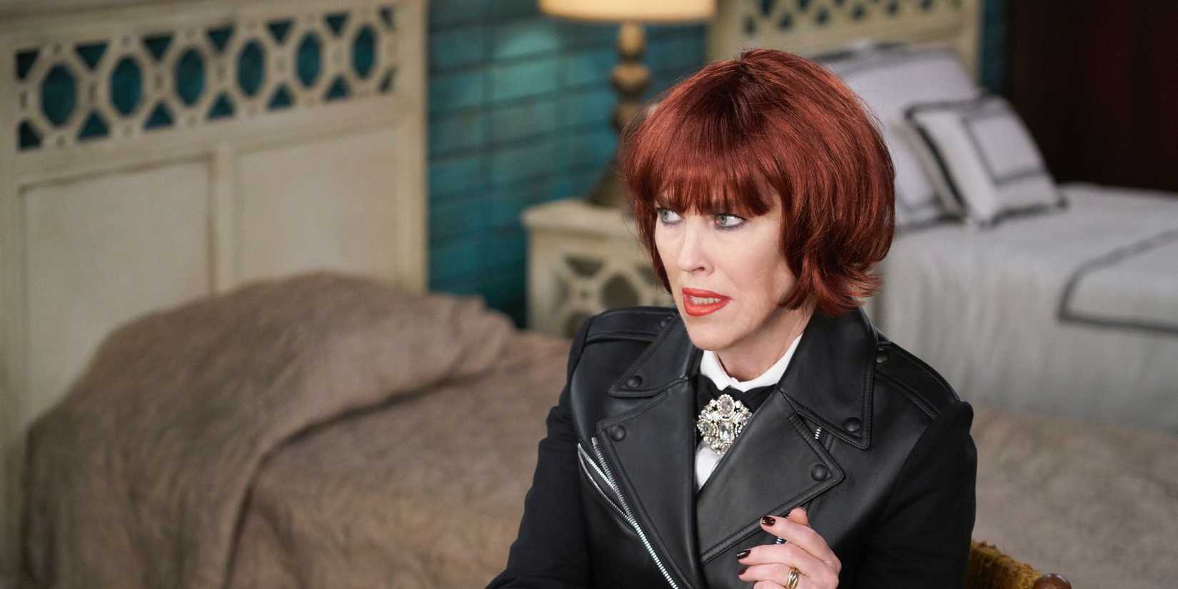 Catherine O'Hara as Moira sitting at a table in the hotel room in a red wig in Schitt's Creek episode The Rollout.