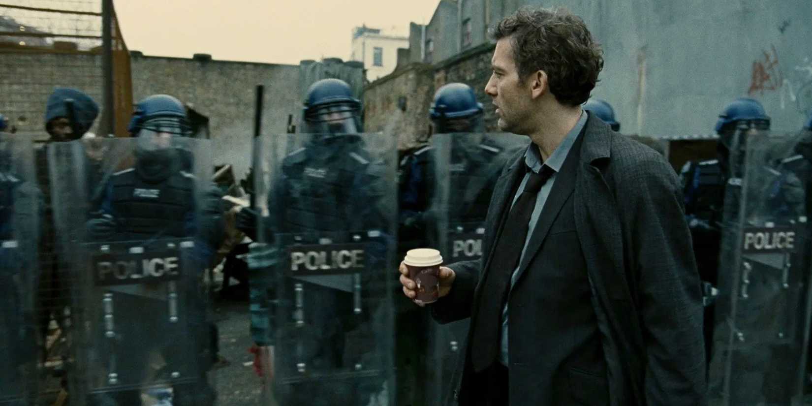 Clive Owen holding a coffee cup while walking past a line of police in Children of Men