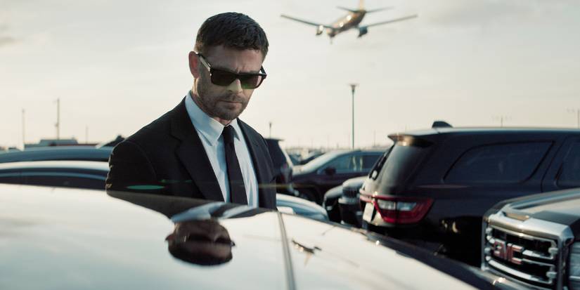 Davis looking down into a car window, wearing sunglasses, as a plane lands in the background.