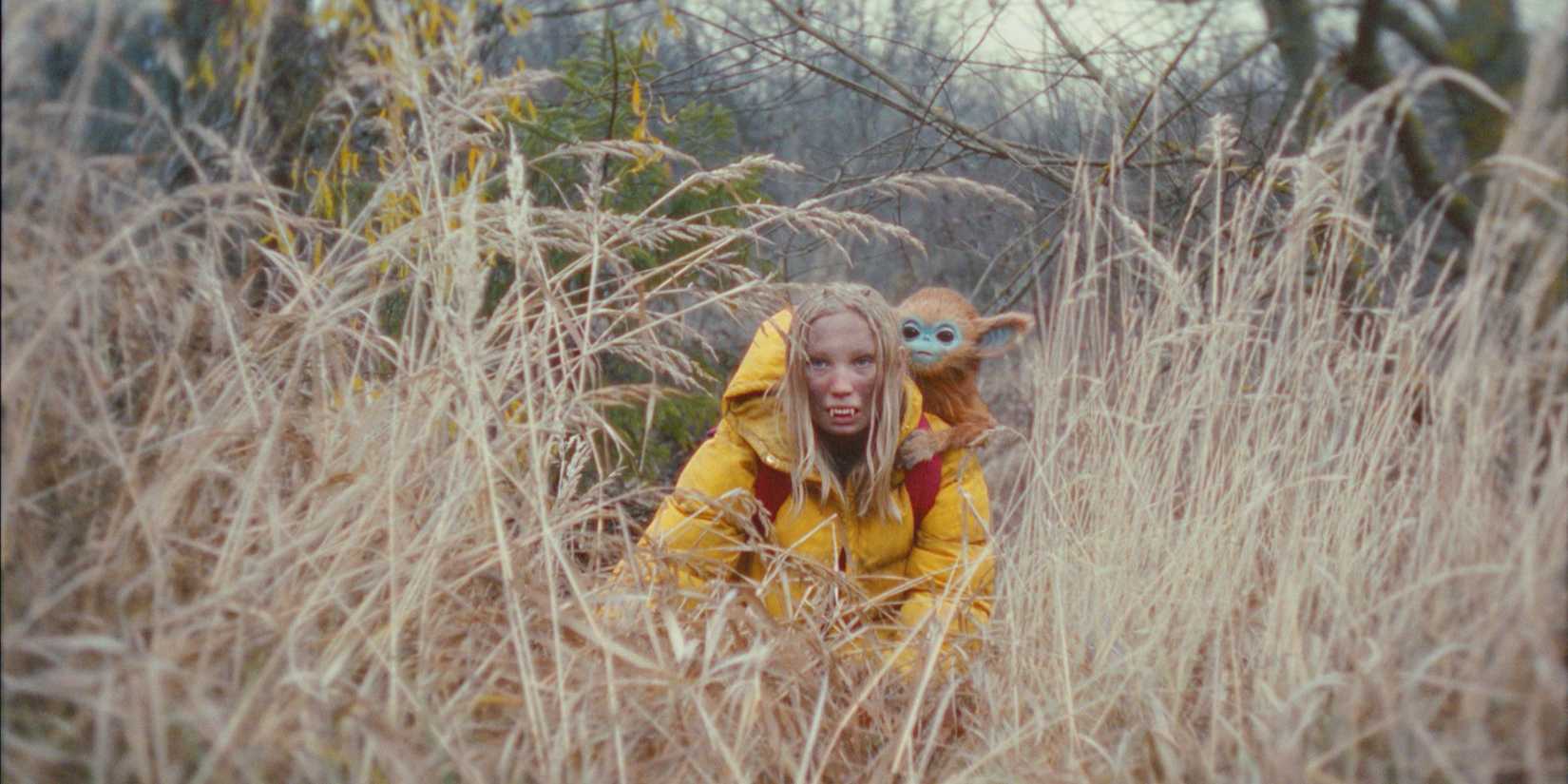 Helena Zengel as Yuri wears fake fanged teeth and crouches in a filed with a baby creature on her back in The Legend of Ochi
