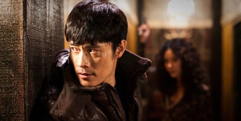 A man hides and looks around a corner with a woman in the distance behind him in I Saw the Devil.