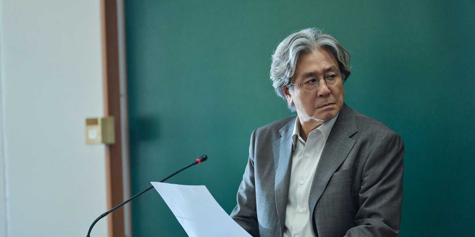 Choi Min-sik standing in front of a microphone stand and holding papers in Notes from the Last Row