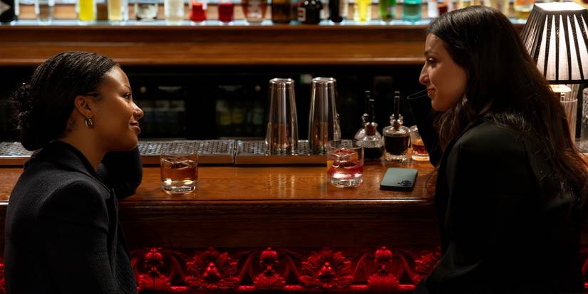 Myha'la as Harper sitting at a bar and smiling at Marisa Abela as Yasmin in Industry Season 4