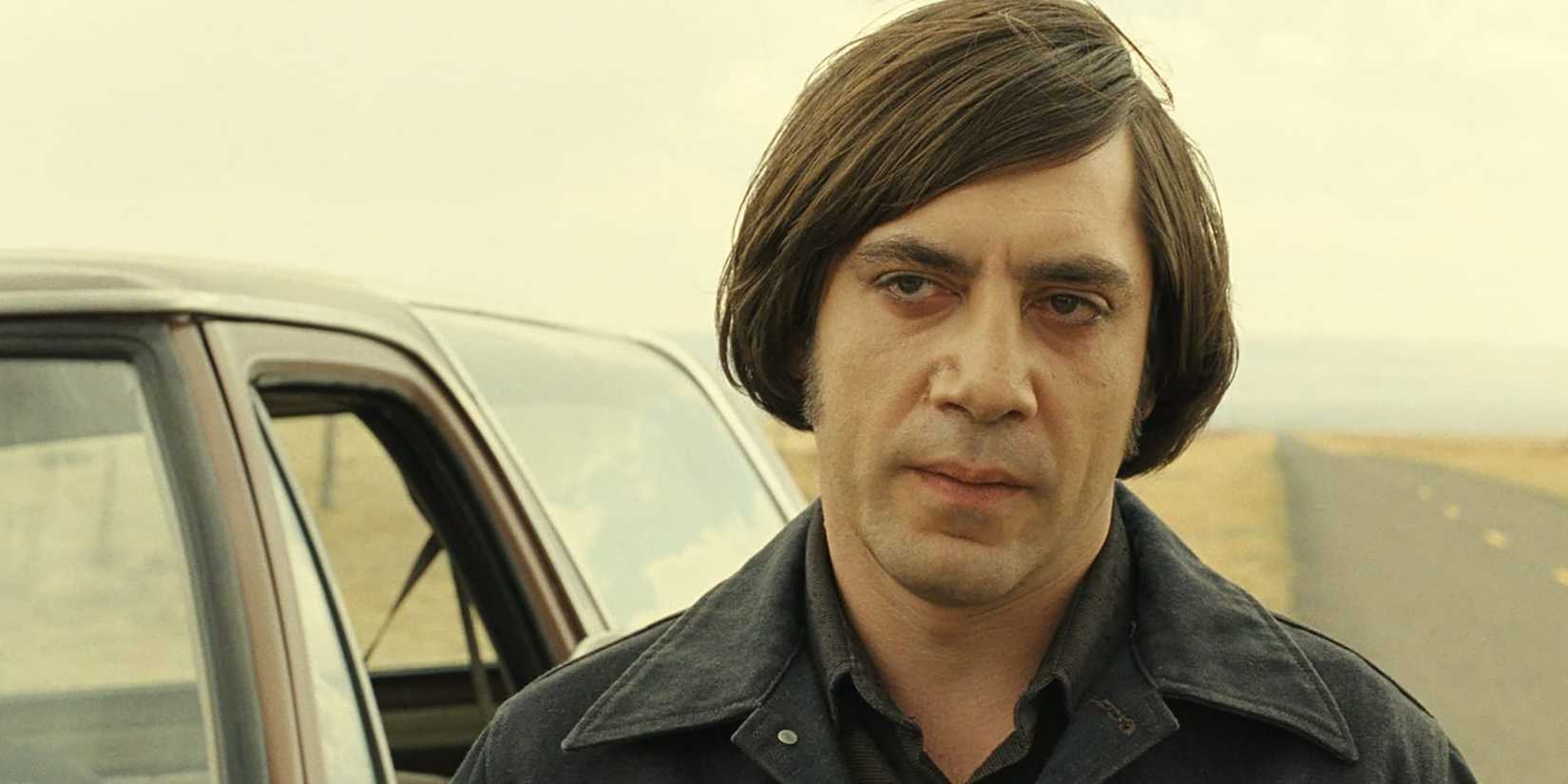Javier Bardem as Anton Chigurh standing near a car in the desert in No Country for Old Men.