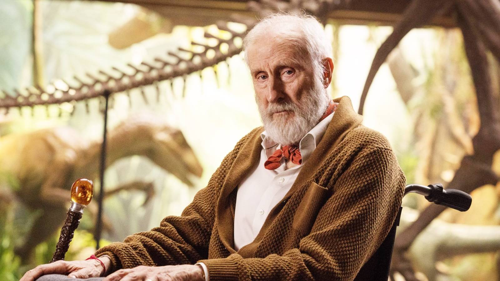 James Cromwell looks at the camera while in a wheelchair in the lobby of Jurassic Park