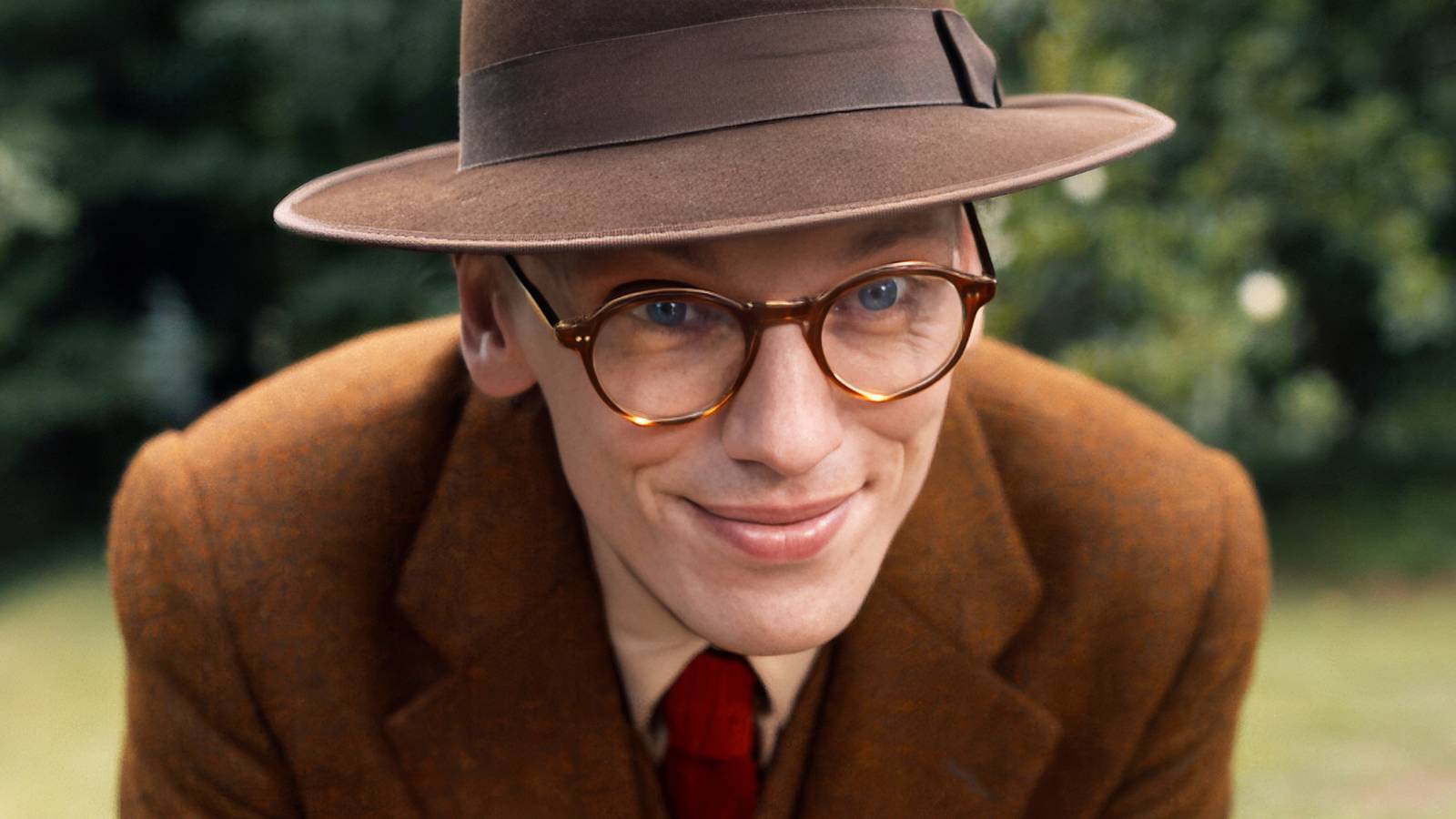 Jamie Campbell Bower in a hat and glasses as Mr. Whatsit, bends over to talk to someone off-camera in Stranger Things.