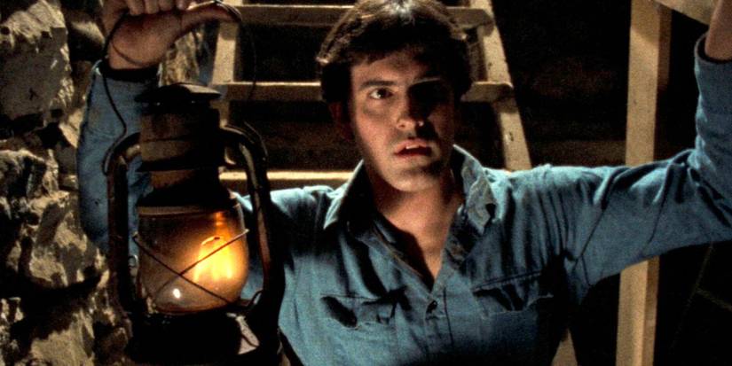A person holds a lamp in a dark cellar and looks wary in The Evil Dead.