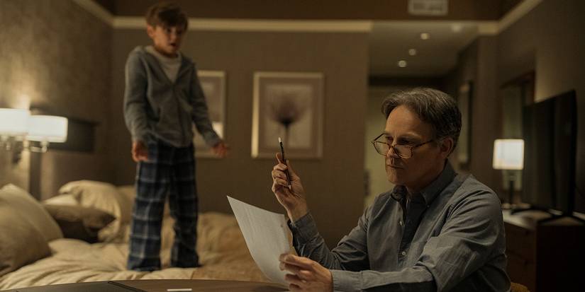 The Father looking at papers in a hotel room as the son stands on the bed in The Night Agent.