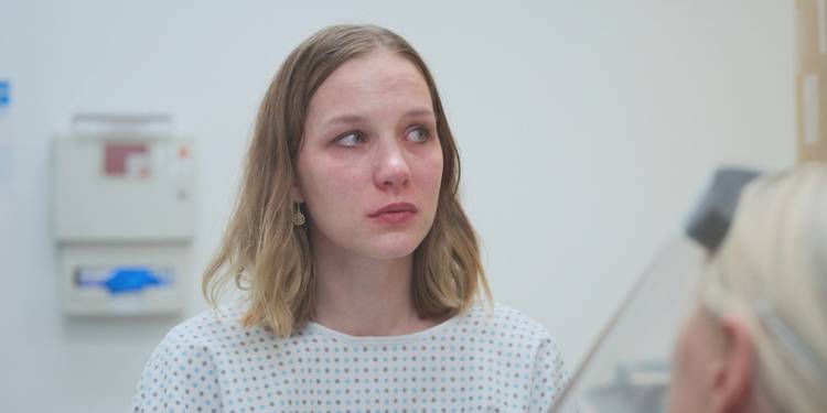 Ilana Miller on the verge of tears while wearing a hospital gown in The Pitt Season 2 Episode 7