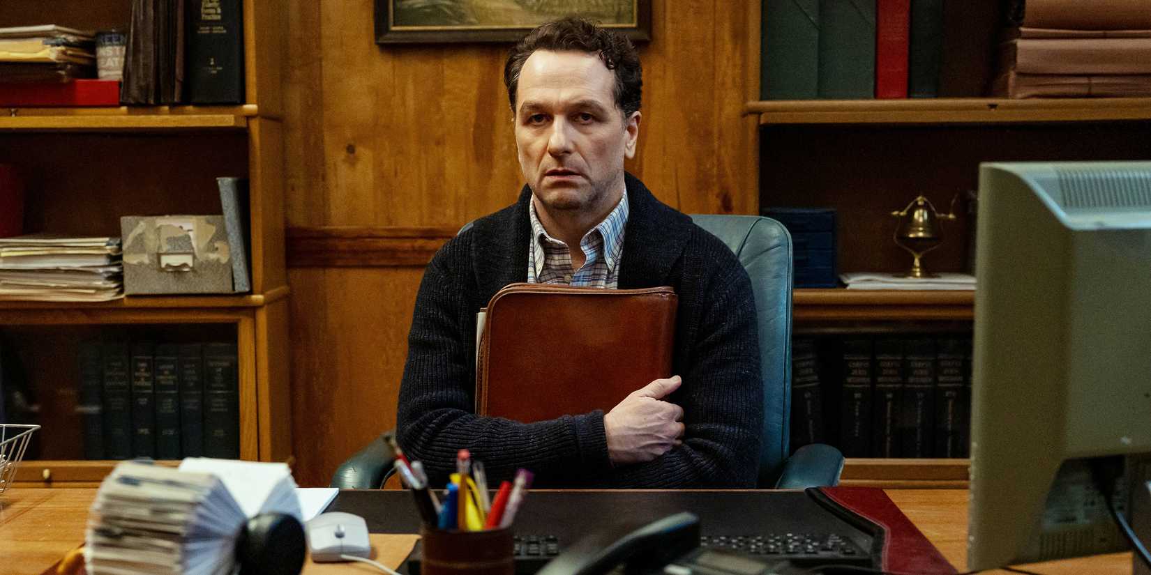 Matthew Rhys as Tom sitting at his office desk while hugging a folder to his chest in Widow's Bay