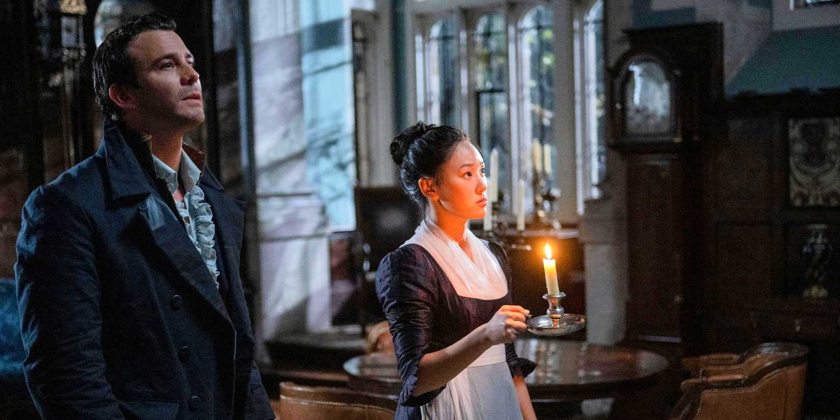 Yerin Ha holds a candle and stands with Luke Thompson in a dark room, both looking in the same direction in Bridgerton.