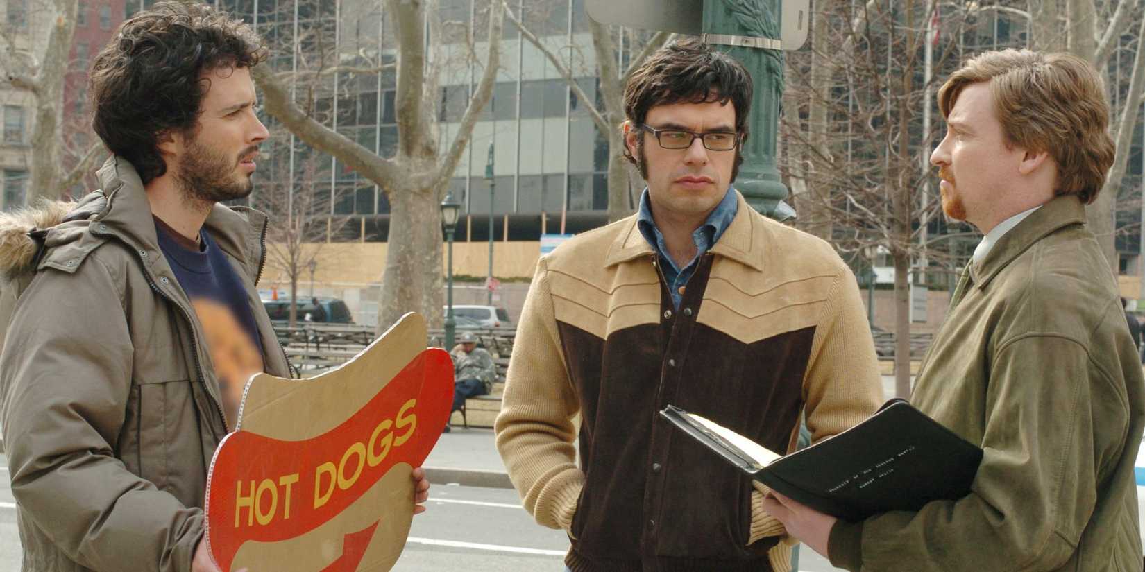 Bret McKenzie, Jemaine Clement, and Rhys Darby talk outside on a street while Bret holds a sign that says "hot dogs" in Flight of the Conchords