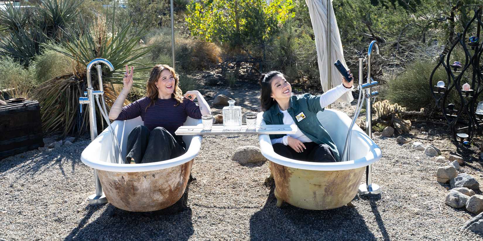 D'Arcy Carden and Sherry Cola smiling and taking a picture in outdoor bathtubs in Wild Vacation Rentals