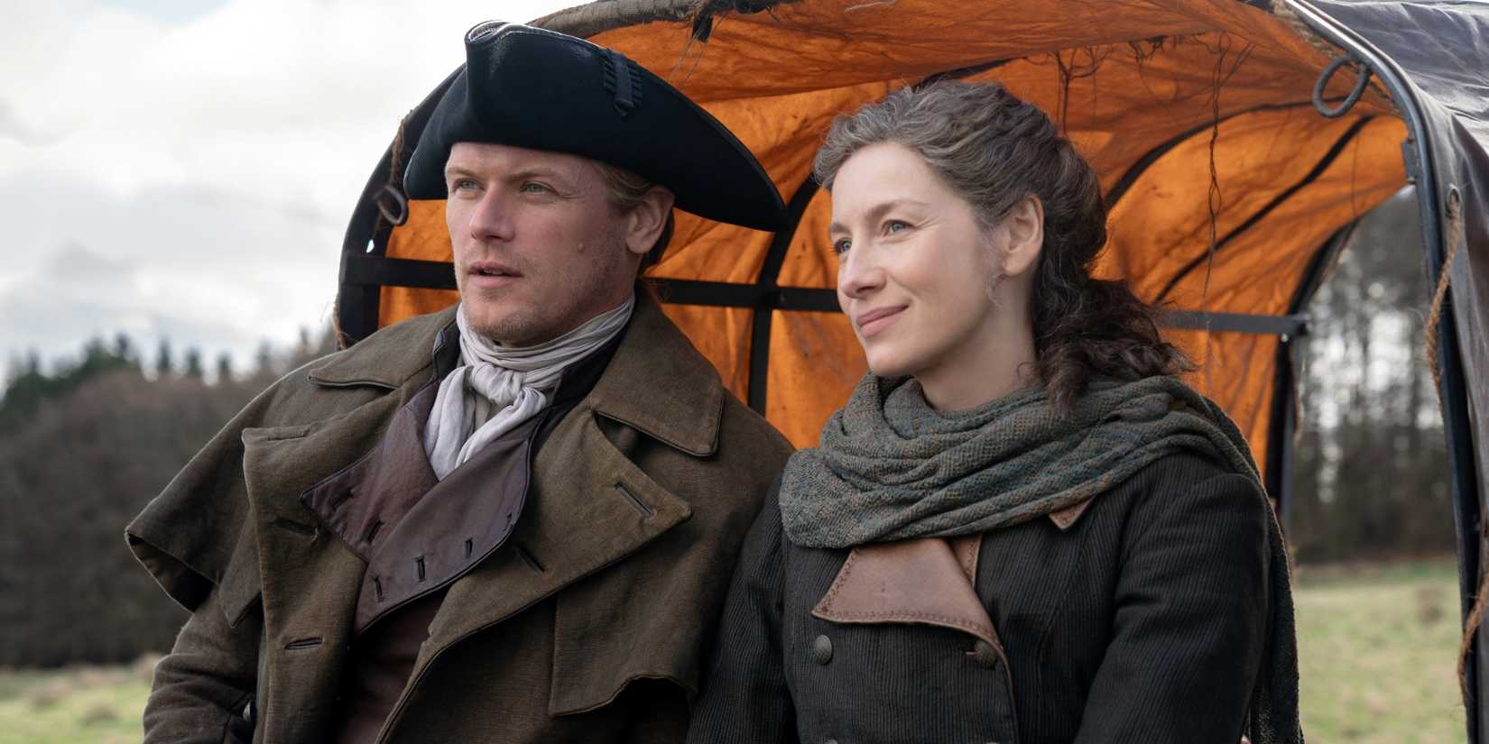 Sam Heughan as Jamie sitting on the back of a wagon next to Caitriona Balfe as Claire in Outlander Season 8