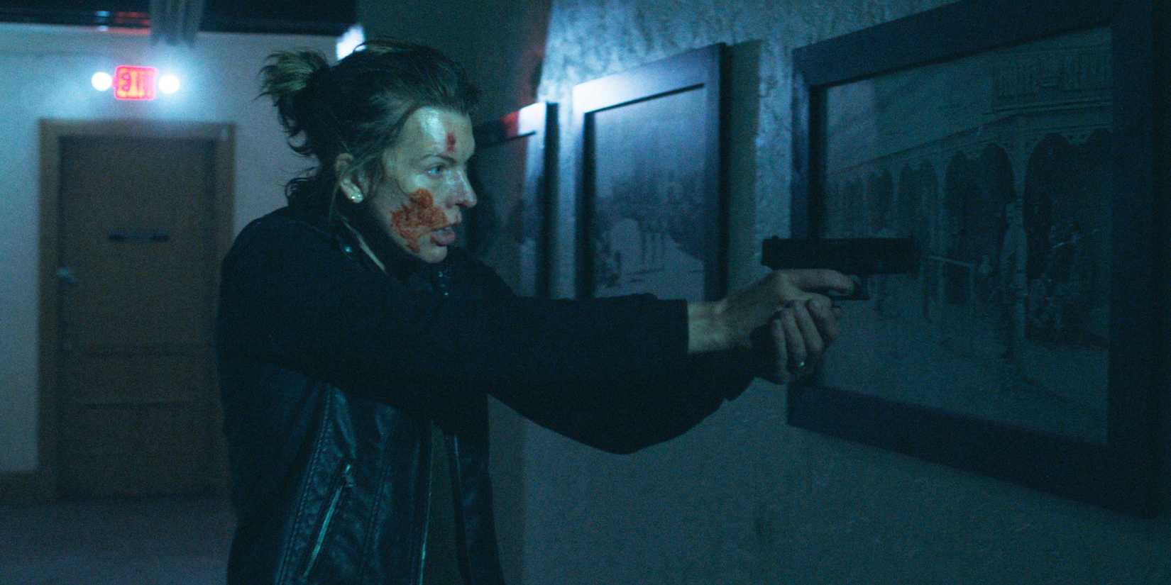 Nikki aims a gun in a dark hallway, with blood on her face.