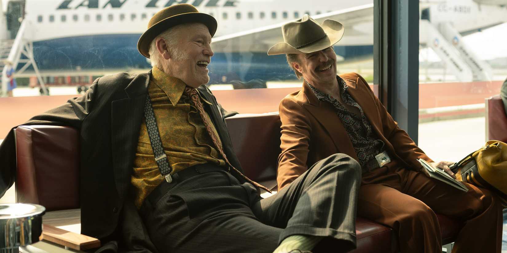 John Malkovich and Sam Rockwell sitting on a couch in an airport and laughing in 'Wild Horse Nine'
