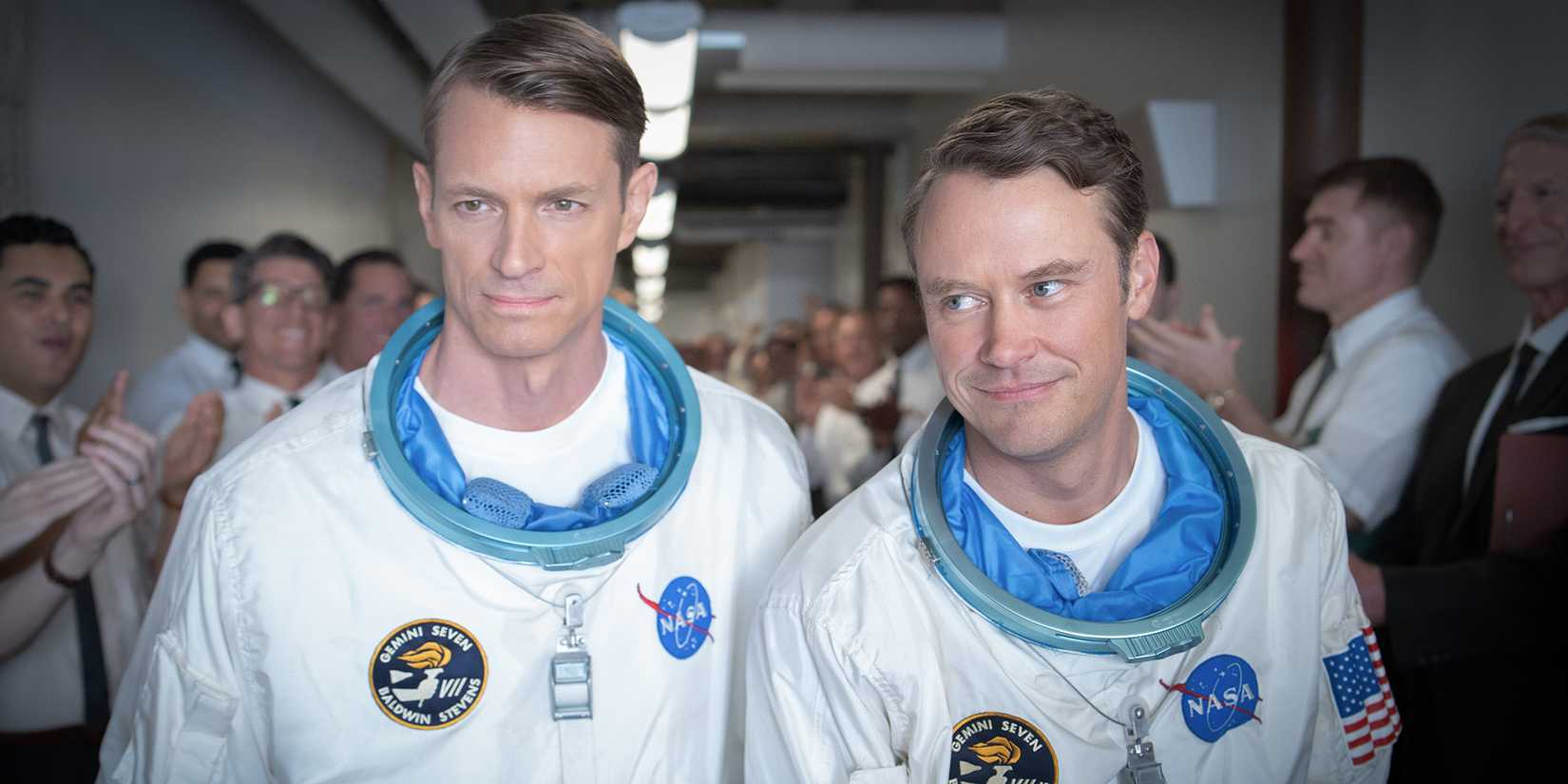Joel Kinnaman and Michael Dorman in their spacesuits walk down a corridor full of a cheering crowd in For All Mankind