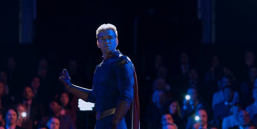 Homelander on stage in the dark as the audience looks out, his hand out to quiet them.