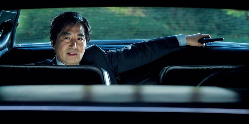 Baek Yoon-sik in the back of a car holding a gun and smiling in The President's Last Bang