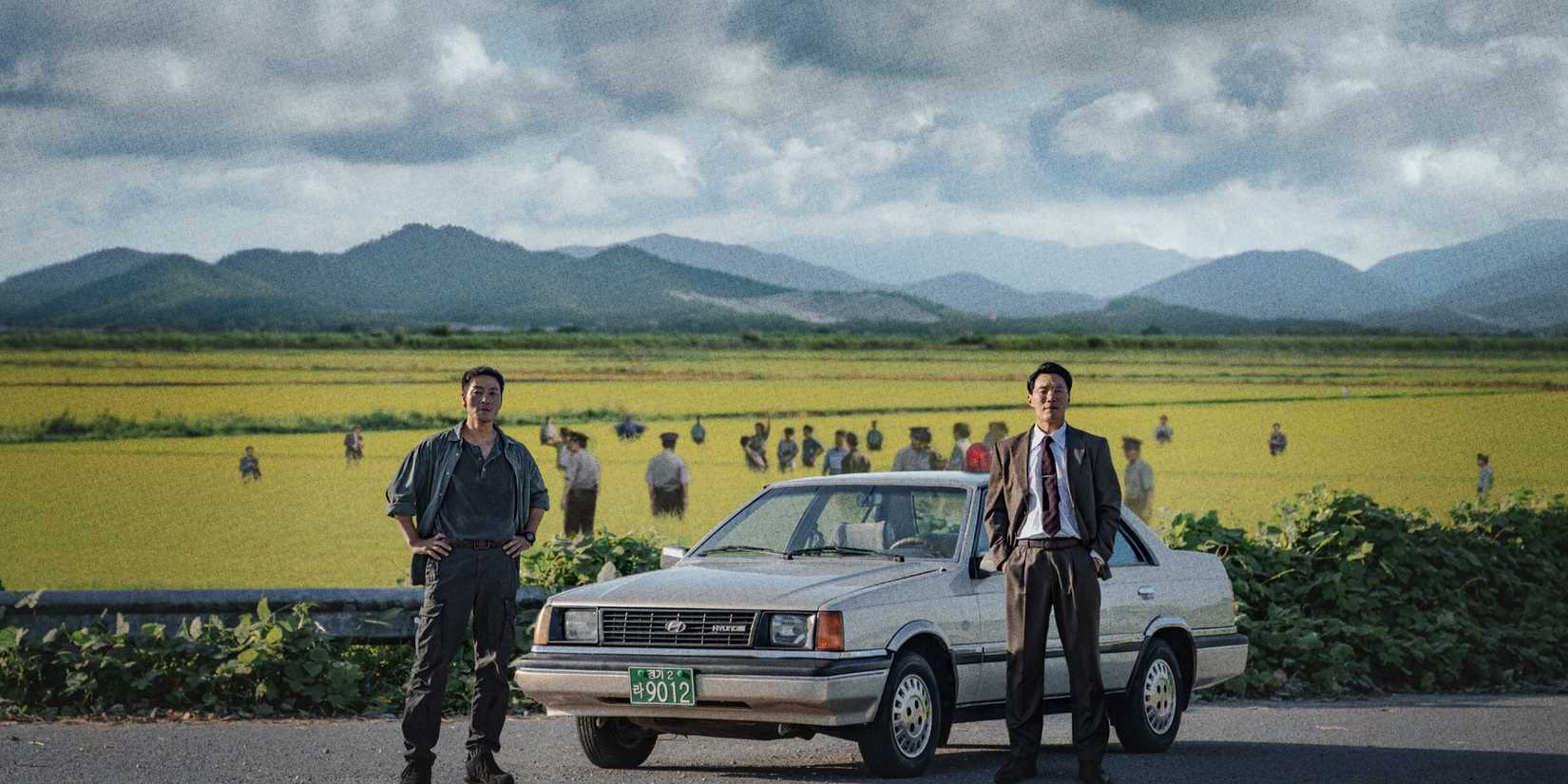 Park Hae-soo and Lee Hee-joon standing next to a vintage car in front of a field full of police officers in The Scarecrow