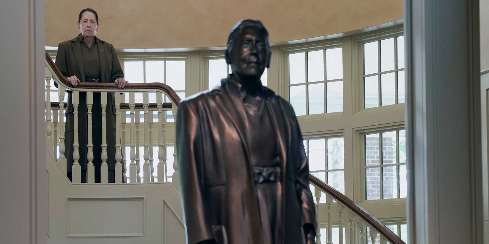 Ann Dowd as Aunt Lydia standing on a stairwell behind her statue in The Testaments