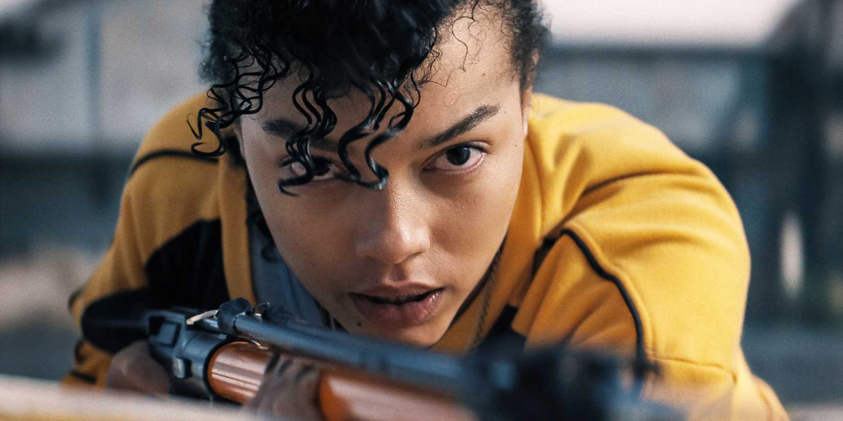 Close-up of Whitney Peak in a yellow jacket, aiming a weapon at something off-camera in Thrash
