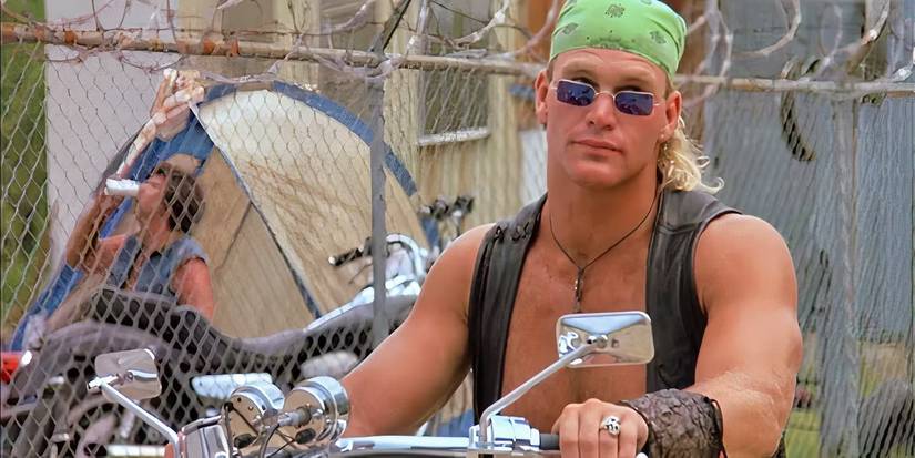 A man in a vest, green bandana, and blue sunglasses is sitting on a motorcycle and holding the wheel while looking over his glasses at something off-screen