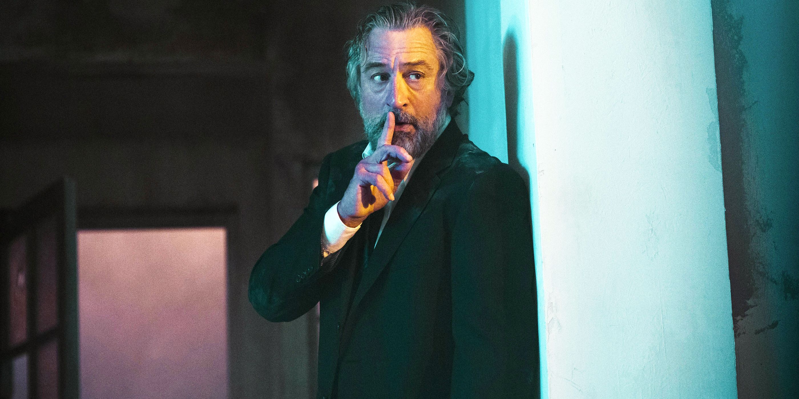 Robert De Niro holding his finger to his mouth as he hides behind a wall in The Family 