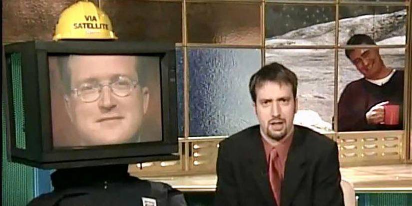 توم جرين يجلس على مكتب يتحدث، ووجه رجل على شاشة تلفزيون بجانبه من برنامج Tom Green Show.