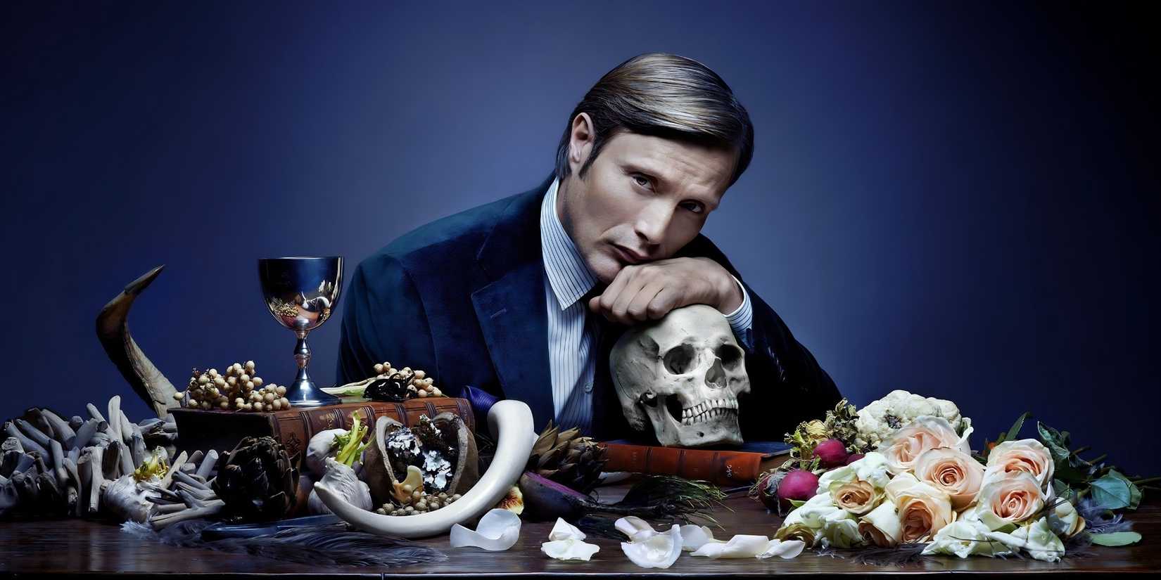 Hannibal Lecter (Mads Mikkelsen) sits at a table, surrounded by bones, flowers and a goblet in a promotional image for Hannibal.